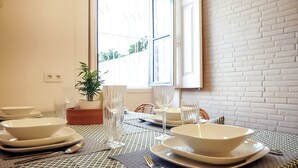 Une table à manger dressée avec des assiettes, des bols, des couverts et des verres, avec une plante en pot et une fenêtre en arrière-plan.