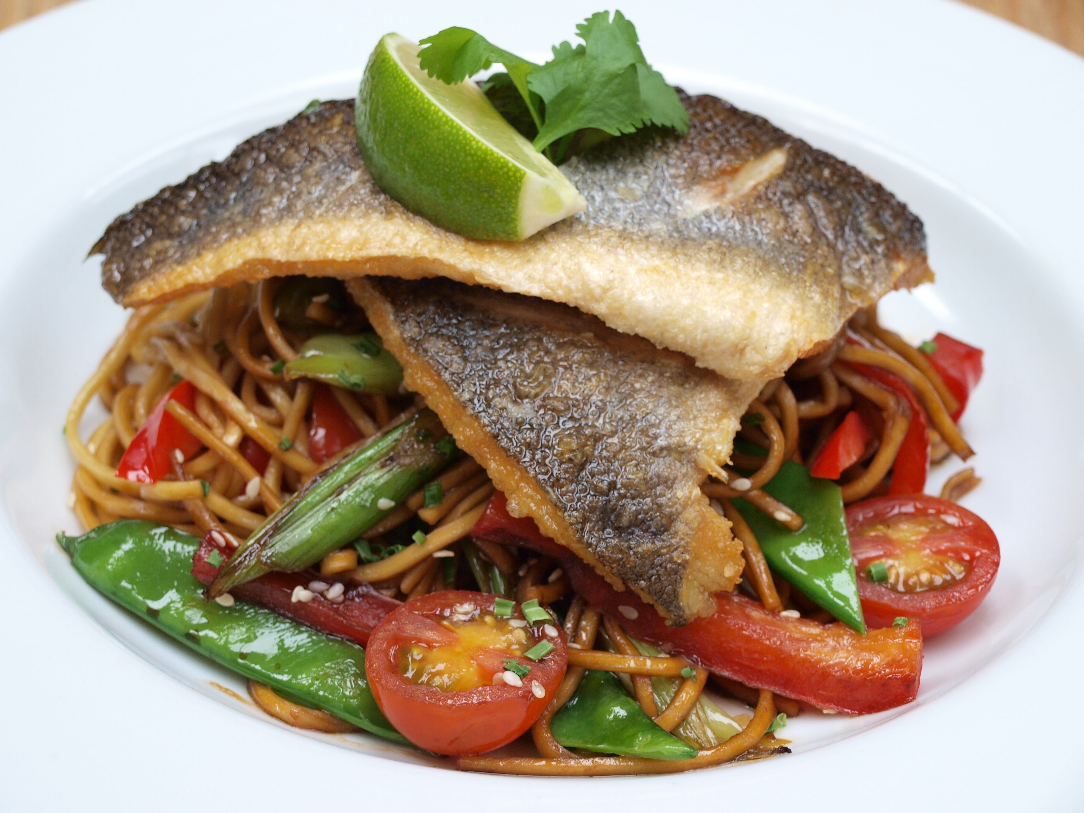 A plate of stir-fried noodles with vegetables and a piece of fish on top.