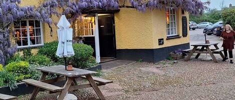 A yellow building with purple wisteria vines, a wooden bench, and a table with a white umbrella.
