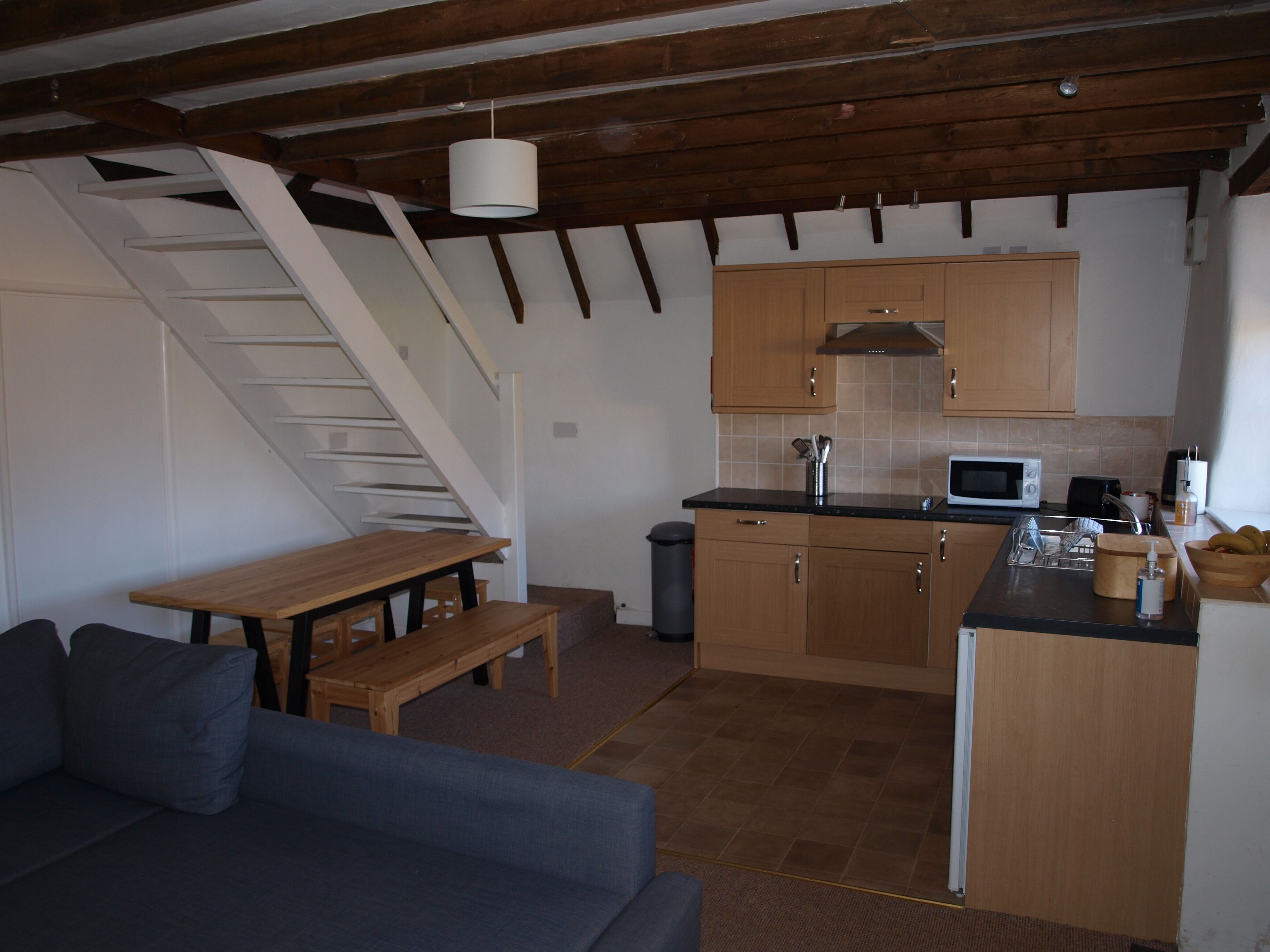 A compact kitchen with wooden cabinets, a microwave, and a sink. There's a wooden bench and a staircase leading upstairs.