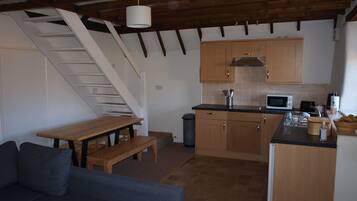 A compact kitchen with wooden cabinets, a microwave, and a sink. There's a wooden bench and a staircase leading upstairs.