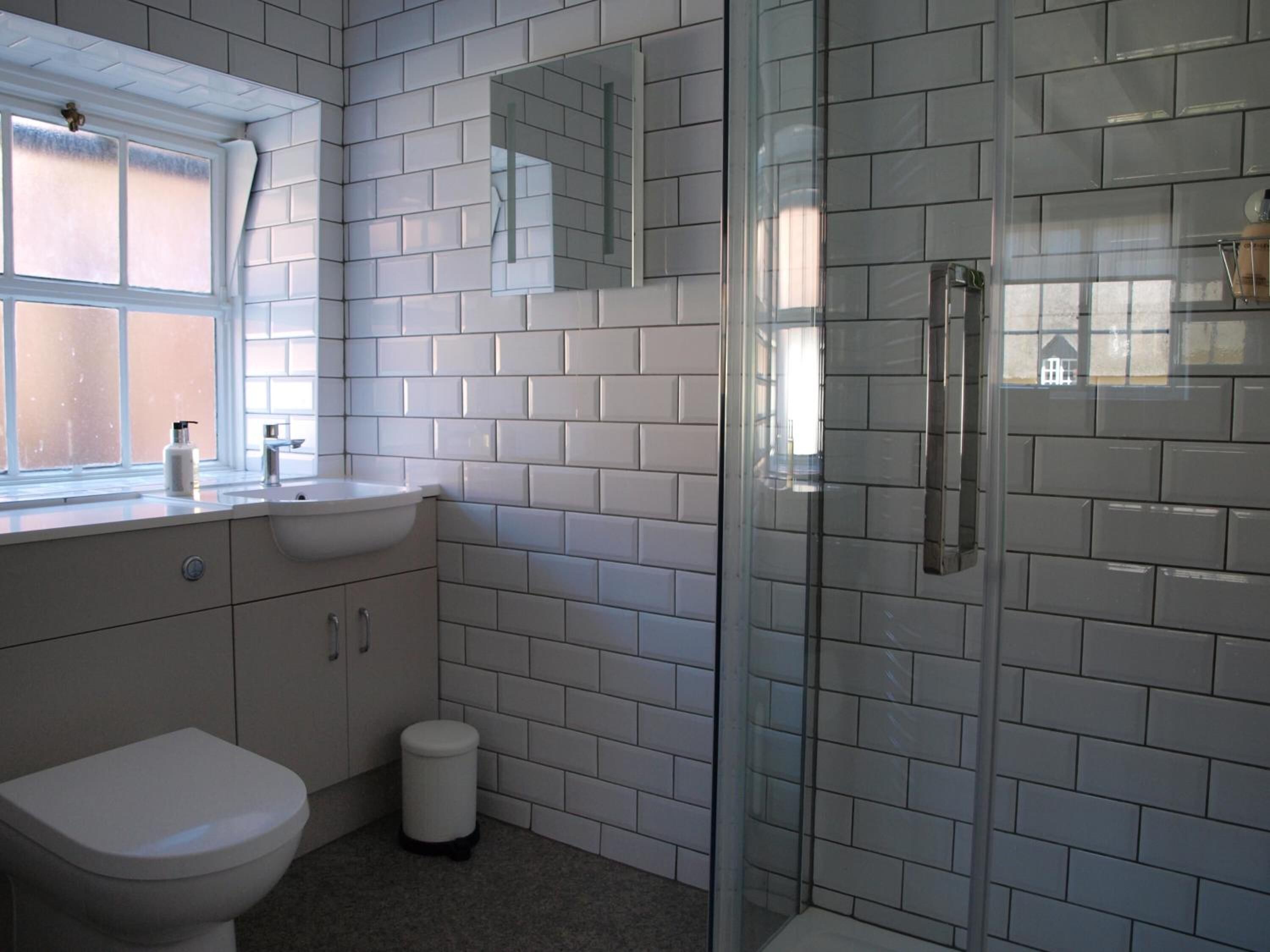 Une salle de bain moderne avec des carreaux de céramique blancs rectangulaires, une douche en verre, des toilettes blanches et une fenêtre qui laisse entrer la lumière naturelle.