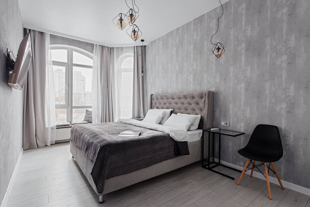 A modern bedroom with a large bed, a side table, a black chair, and a view of the city through large windows.