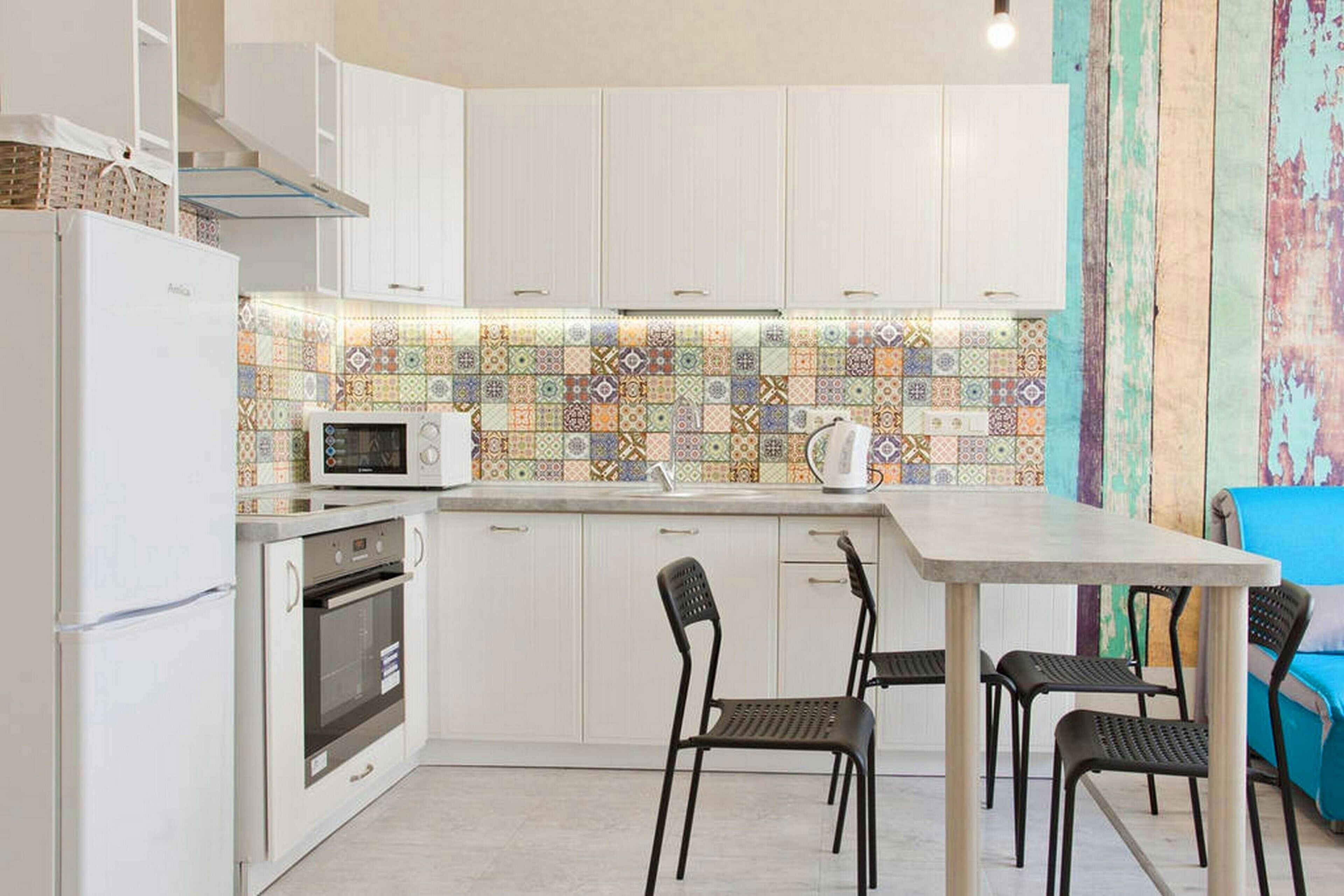 Eine kompakte Küche mit weißen Schränken, einer Mikrowelle, einem Backofen und einer gefliesten Spritzschutzwand. Vier schwarze Stühle stehen um einen kleinen Tisch.