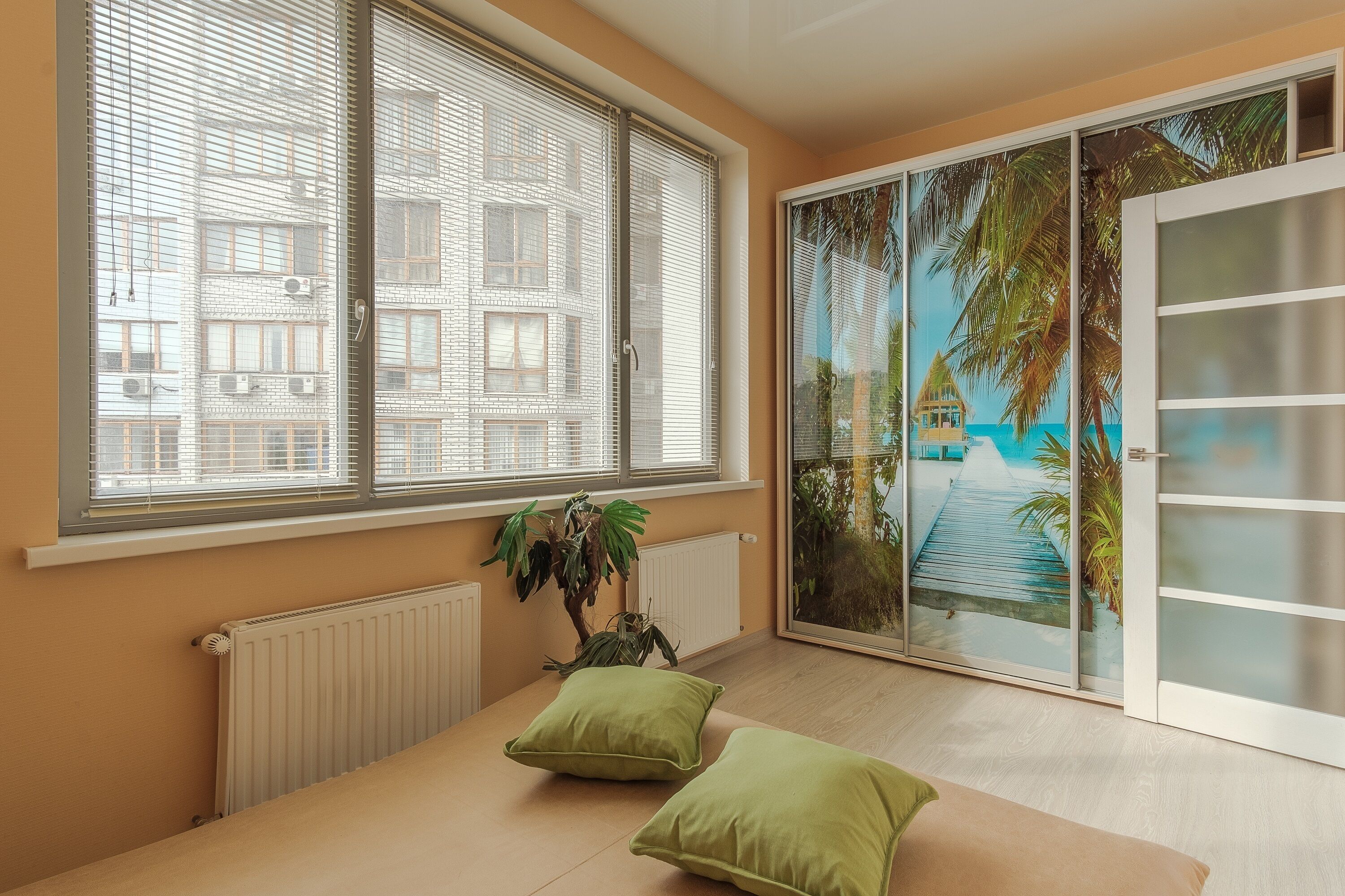A room with a large window, a radiator, a plant, and a view of a beach through a sliding door.
