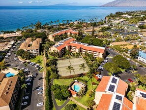 An aerial view of a coastal resort area with multiple buildings, a tennis court, and a swimming pool.