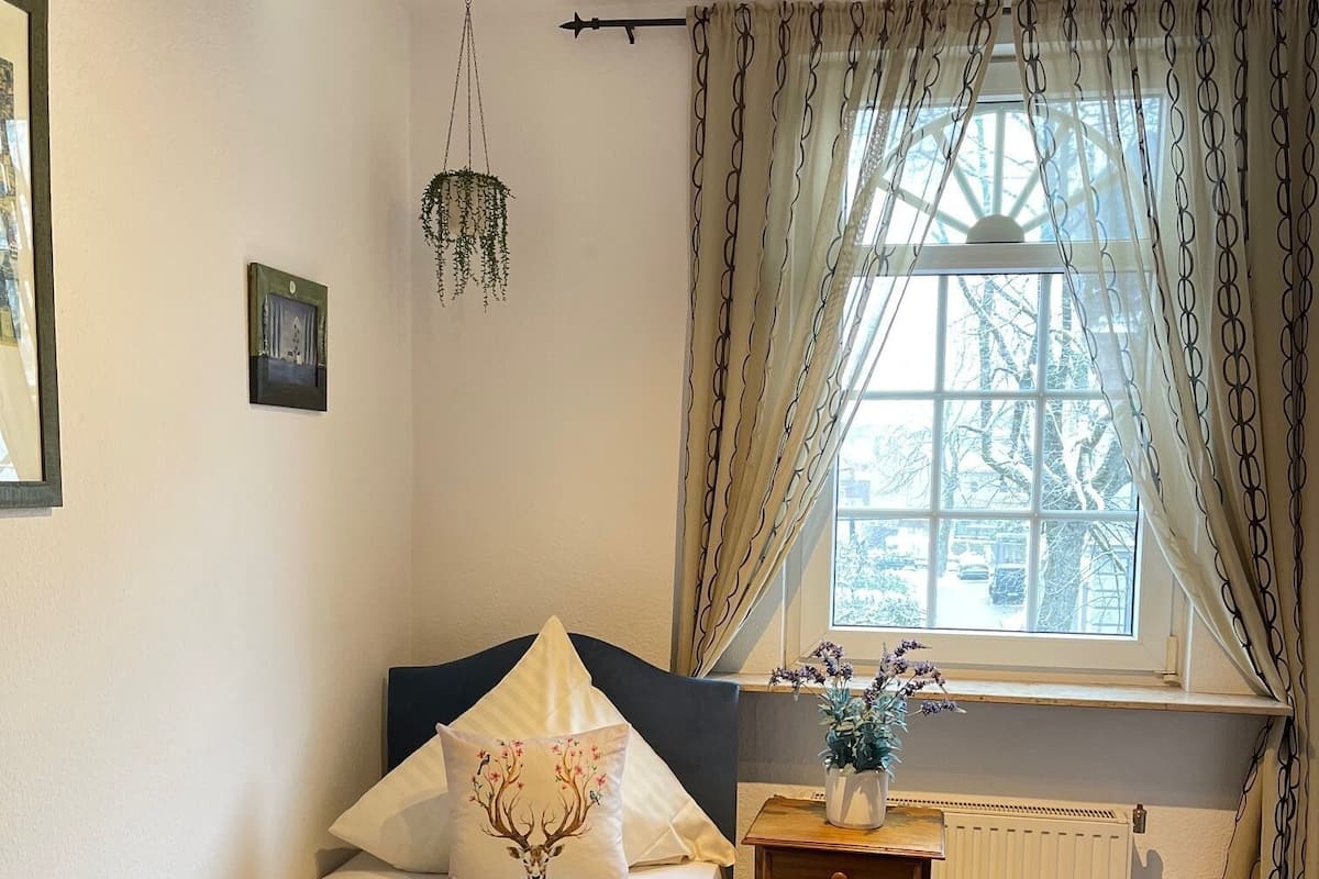 Une chambre à coucher avec un lit, une table de chevet sur laquelle se trouve un vase, une plante suspendue et une fenêtre avec des rideaux.