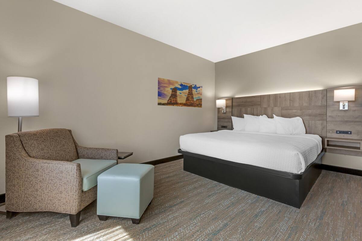 A hotel room with a bed, a chair, a nightstand, a lamp, and a painting on the wall.