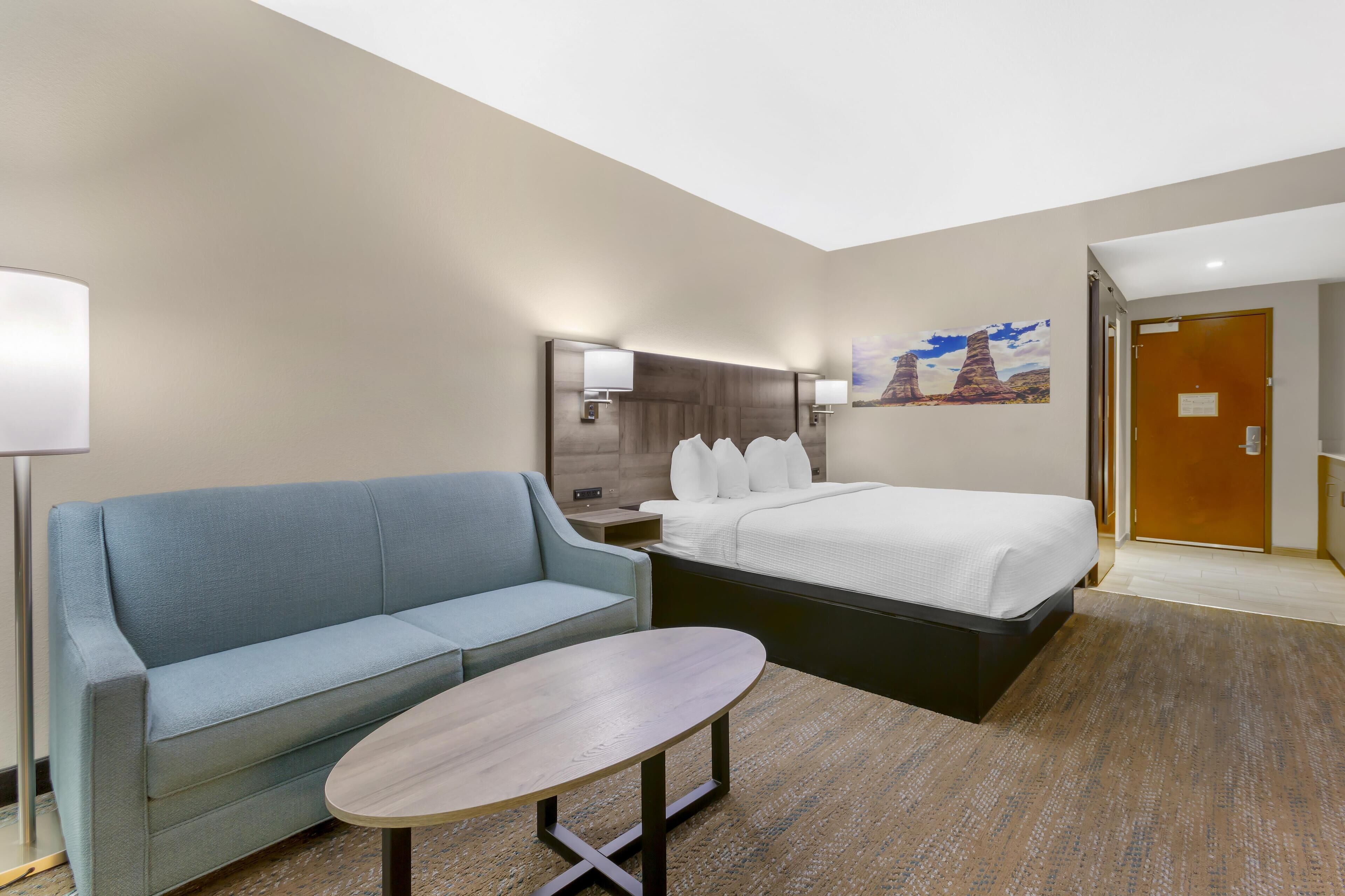 Habitación de hotel con cama, sofá, una mesita, mesita de noche, lámpara y un cuadro en la pared.