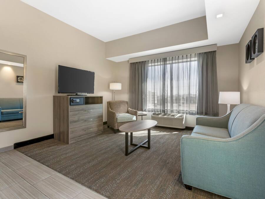 Quarto de hotel com televisão de tela plana, um sofá, uma poltrona, uma mesa de centro e uma janela grande com cortinas.
