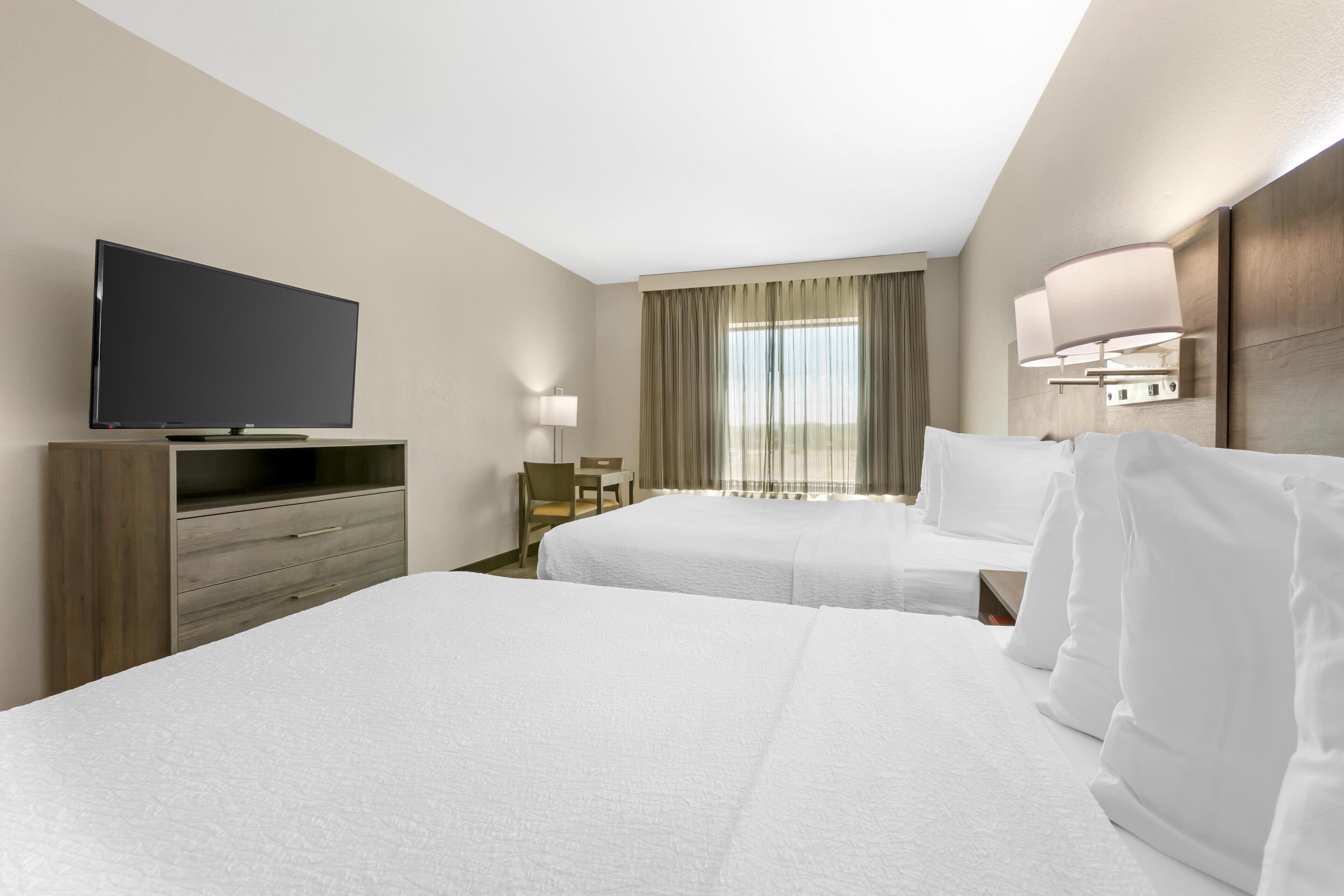 Une chambre d’hôtel avec deux lits, un téléviseur posé sur un meuble en bois, un bureau avec une chaise et une fenêtre avec des rideaux.