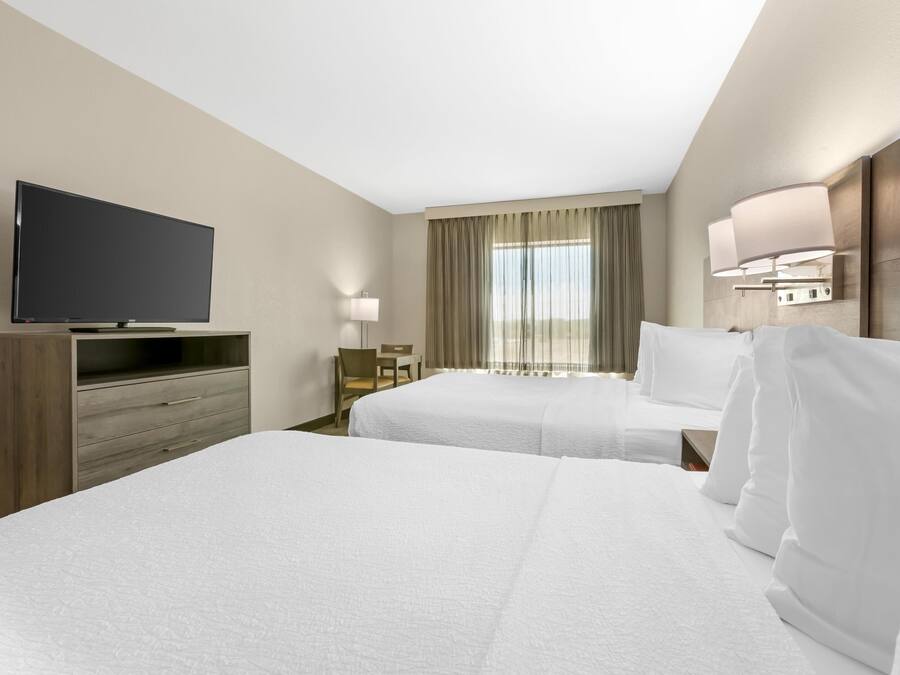 Une chambre d’hôtel avec deux lits, un téléviseur posé sur un meuble en bois, un bureau avec une chaise et une fenêtre avec des rideaux.