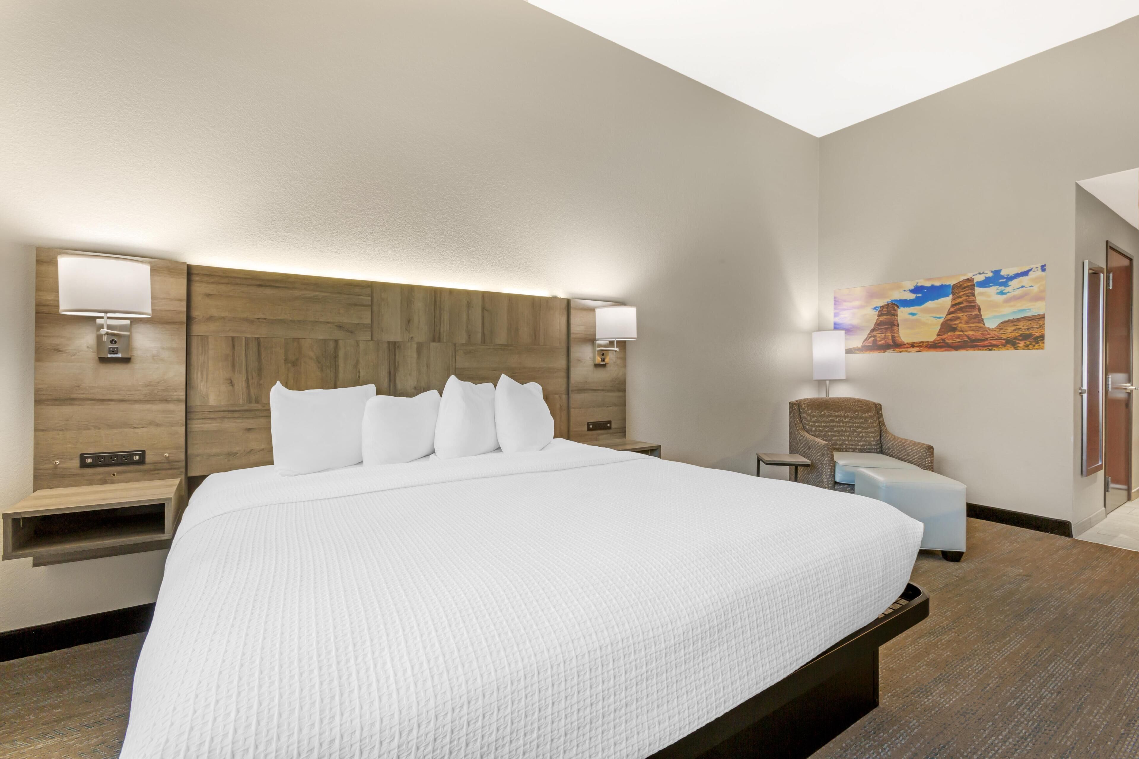 A modern hotel room with a large bed, a wooden headboard, two bedside lamps, a chair, and a painting on the wall.