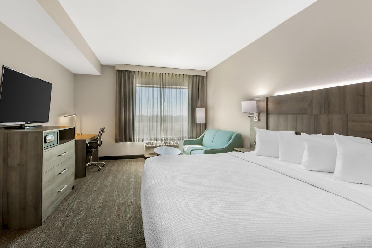 A hotel room with a large bed, a desk with a flat-screen TV, a chair, a small table, and a window with curtains.