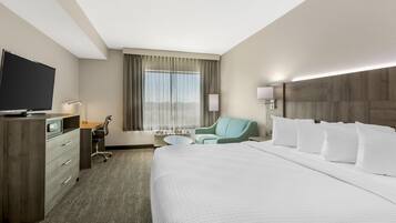 A hotel room with a large bed, a desk with a flat-screen TV, a chair, a small table, and a window with curtains.