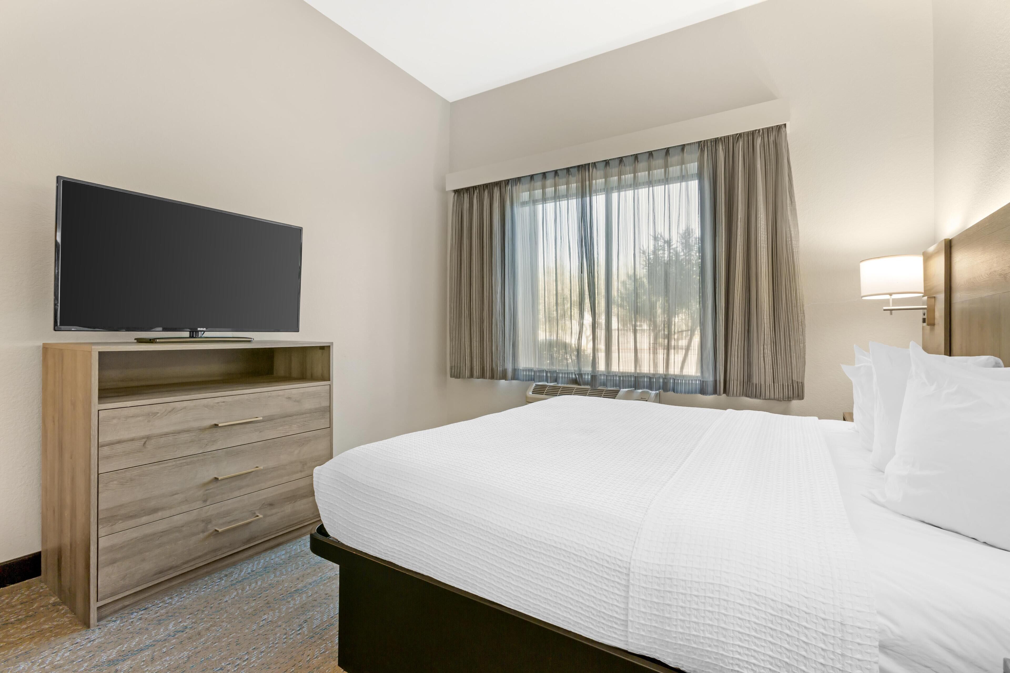 Ein Hotelzimmer mit einem Bett, einem Fernseher auf einer Holzkommode, einem Fenster mit Vorhängen und einer Wandlampe.