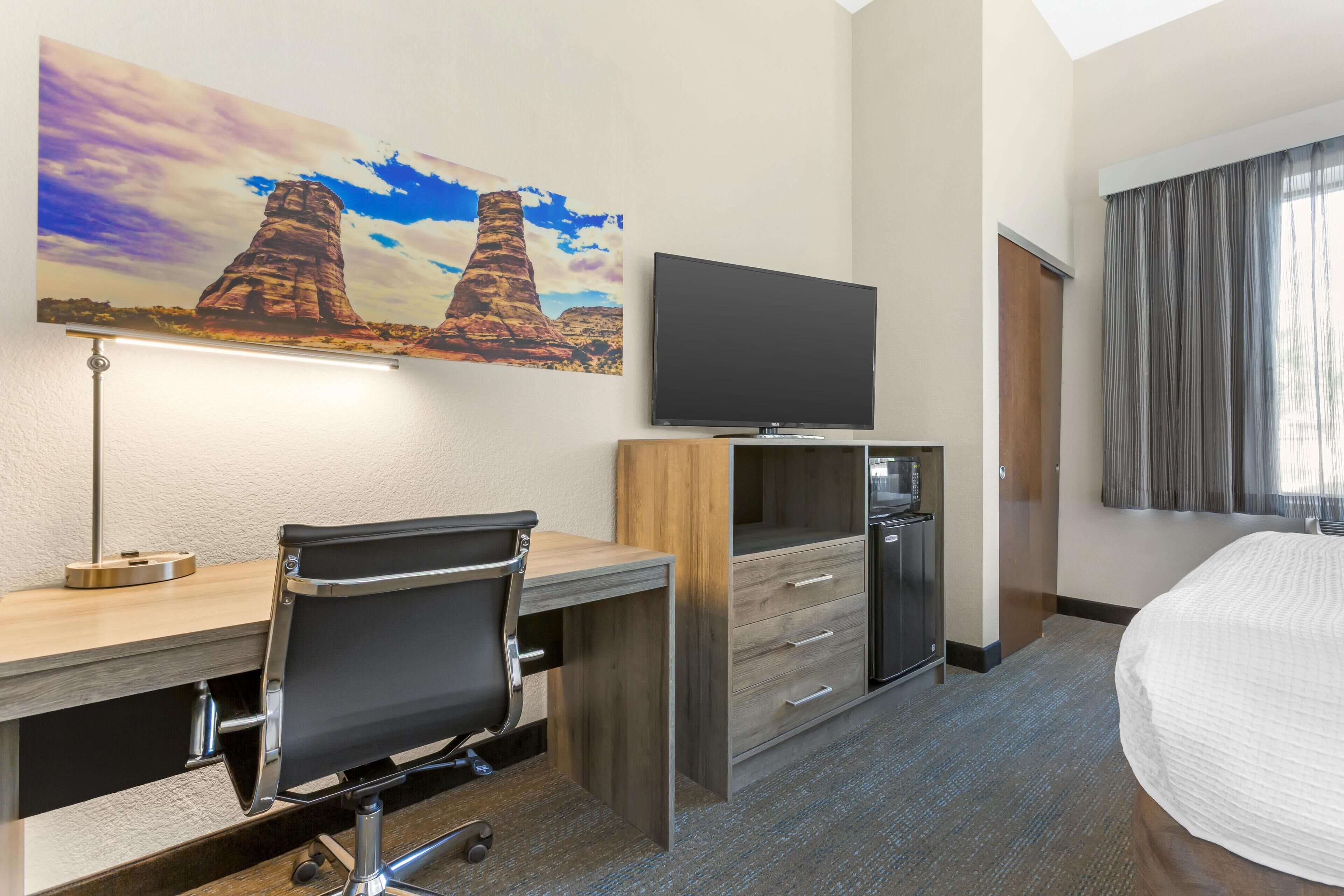A hotel room with a desk, chair, television, bed, and a painting of rock formations.
