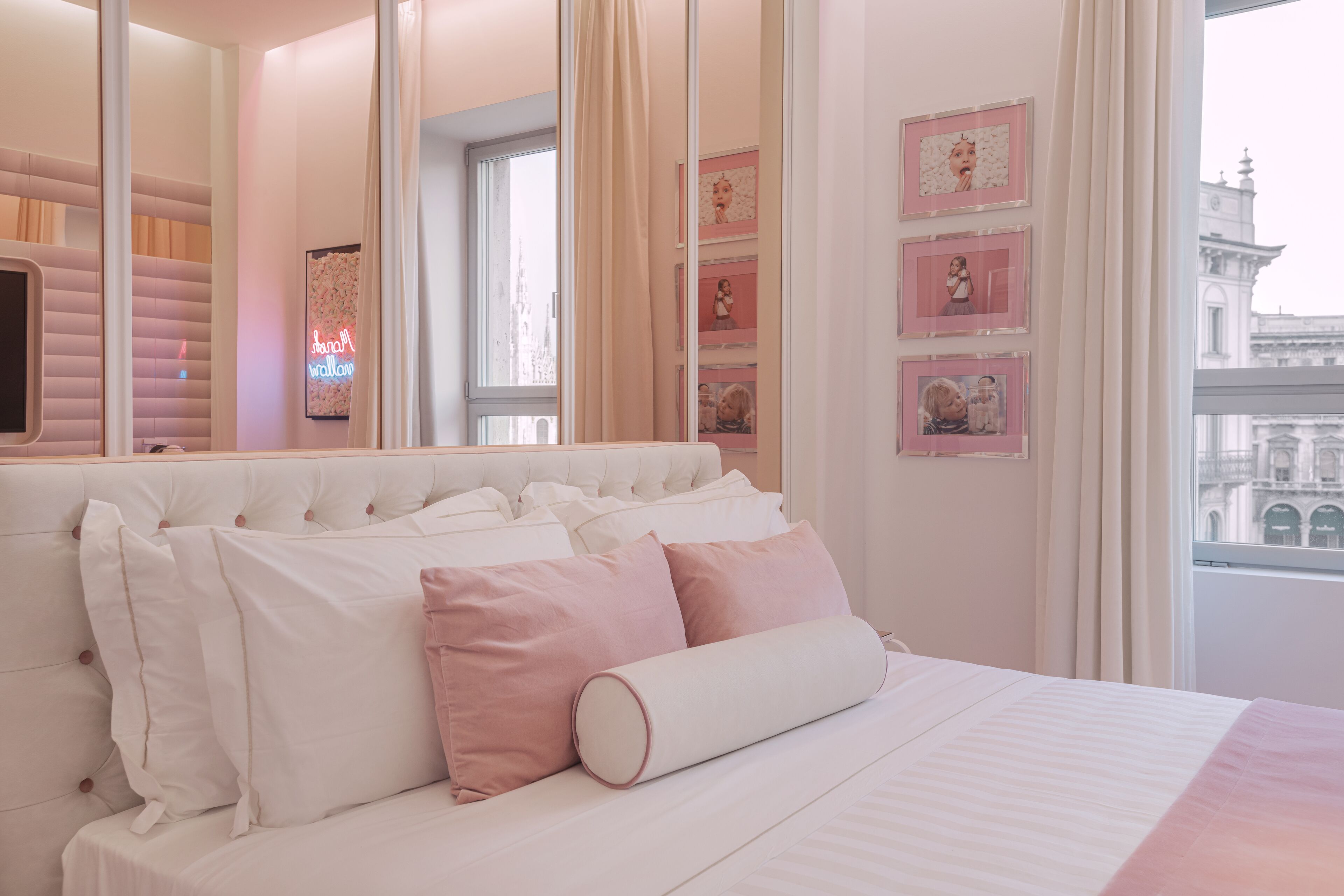 A neatly arranged bedroom with a large bed, a window with a view of a building, and a neon sign on the wall.