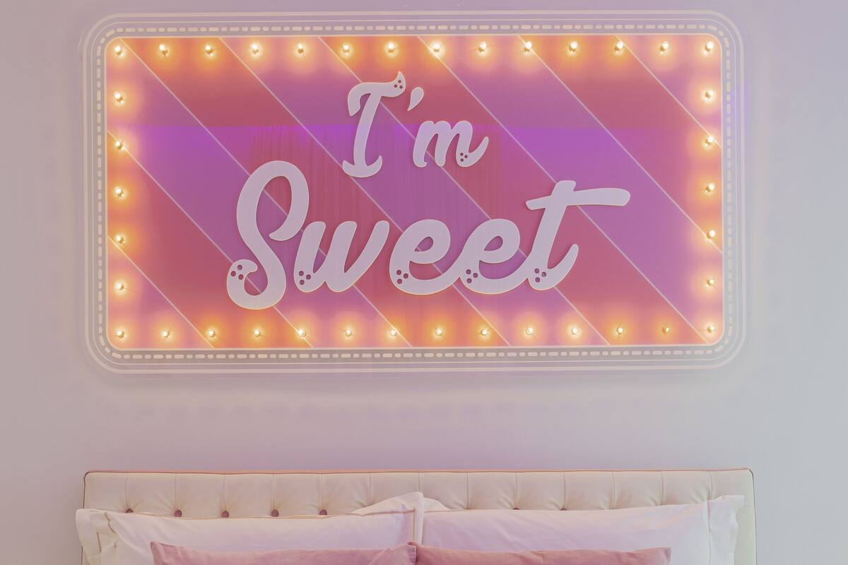 A bedroom with a bed, two pillows, and a neon sign on the wall that reads "I'm Sweet".