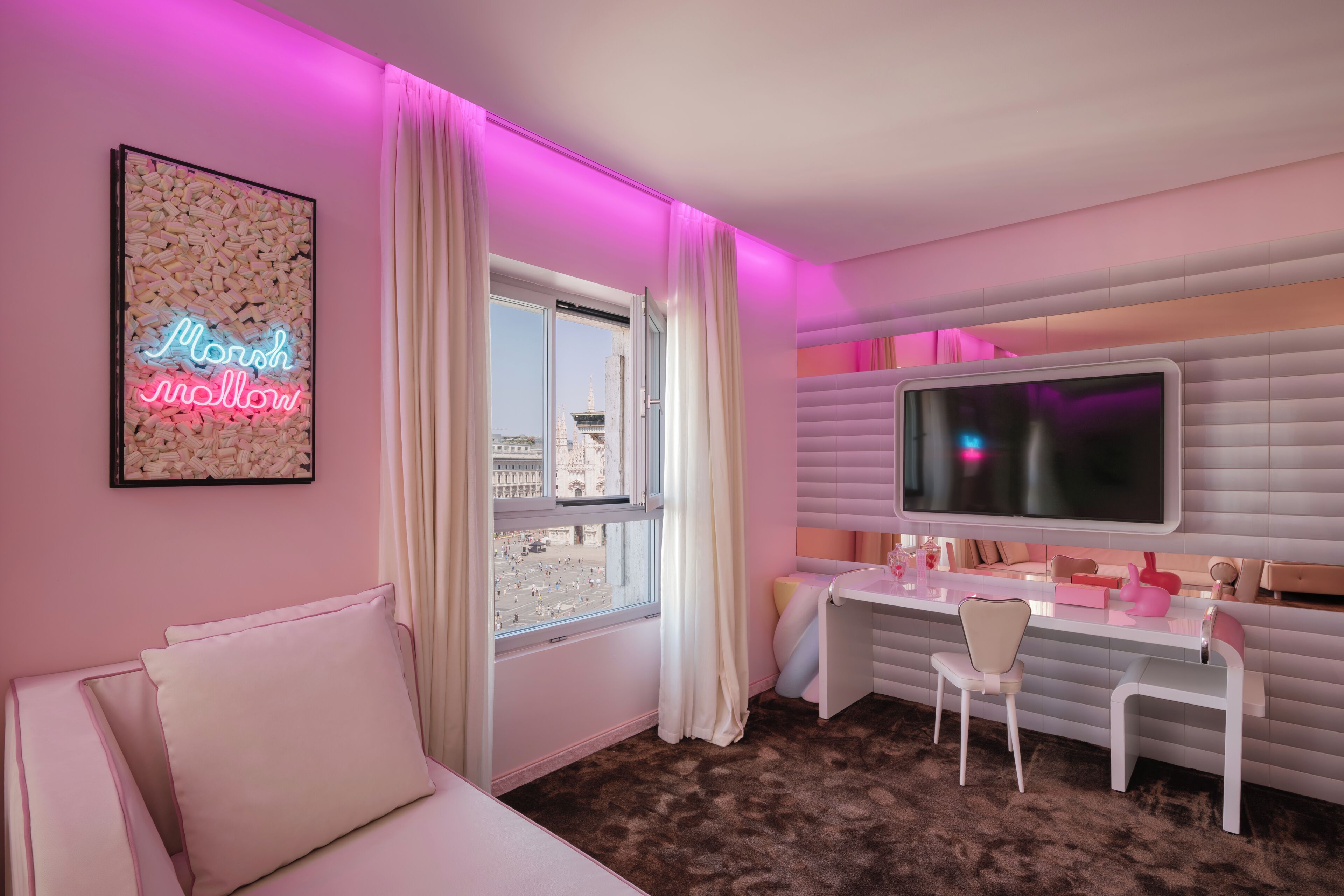 Habitació de color rosa amb un llit, un escriptori, un televisor i una finestra amb cortines.