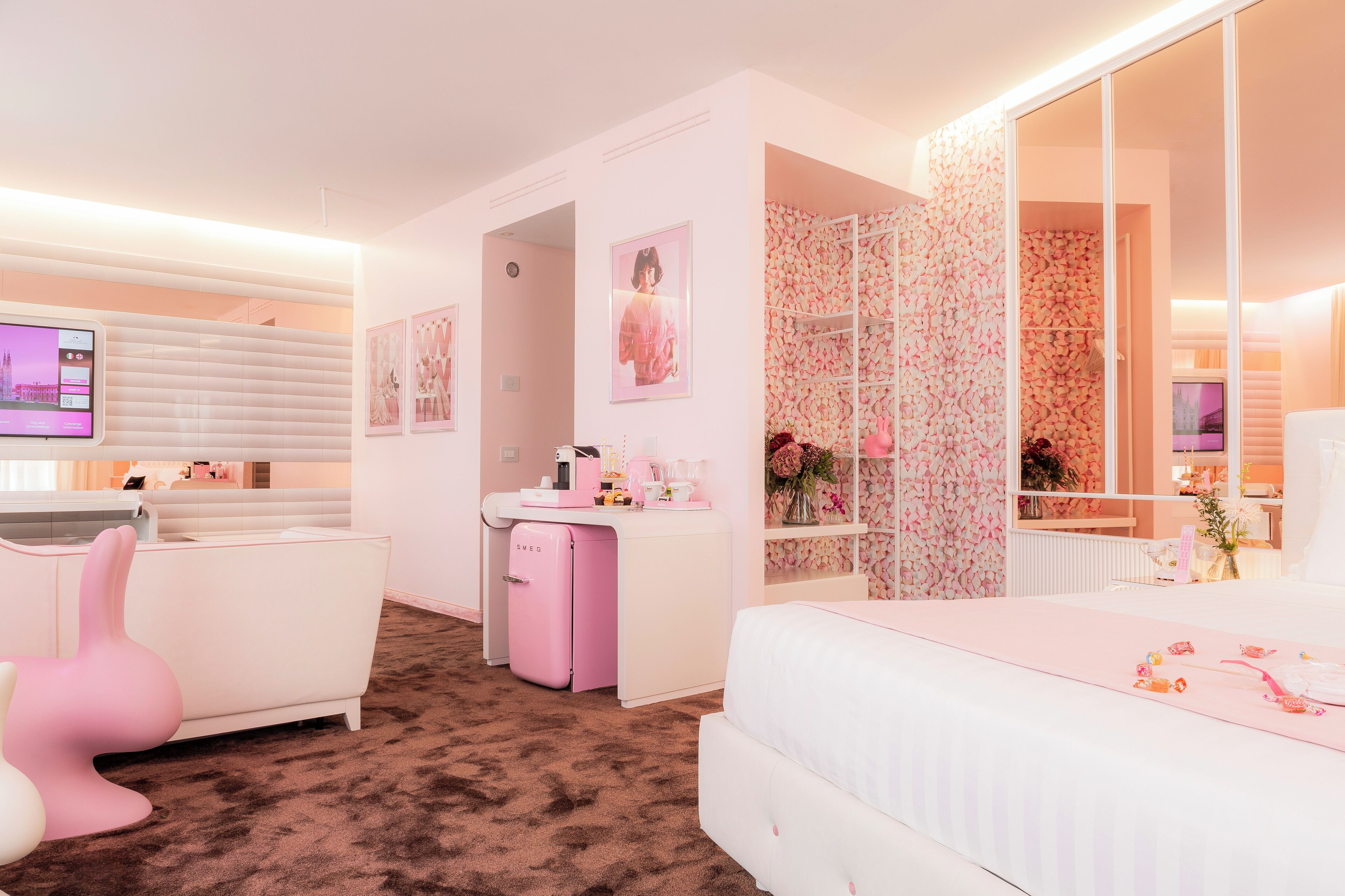 Une chambre décorée dans les tons de rose, avec un lit blanc, une chaise rose et un petit réfrigérateur rose.