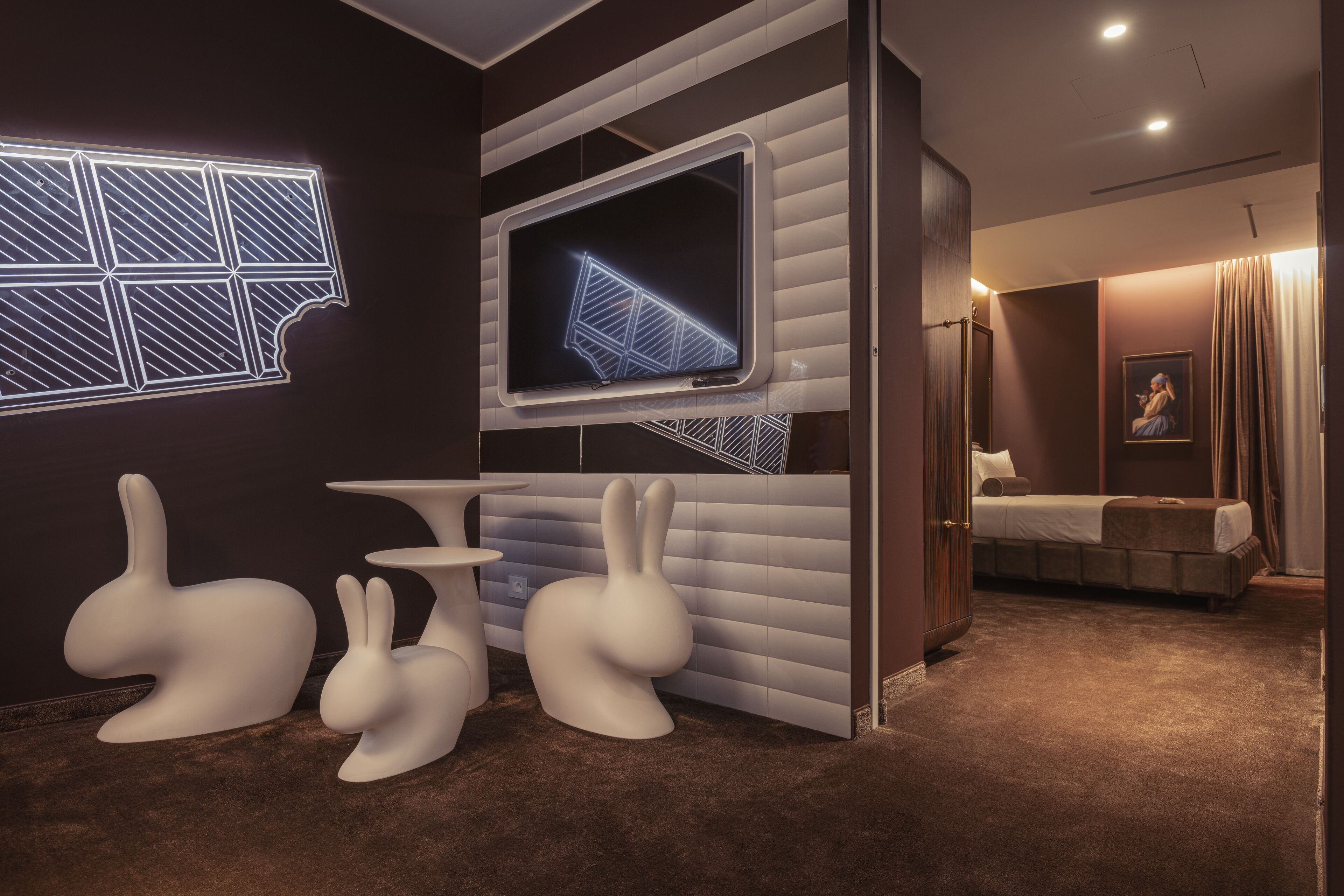 A modern hotel room with a large flat-screen TV mounted on the wall, a bed with a headboard, a small table, and two white rabbit-shaped stools.