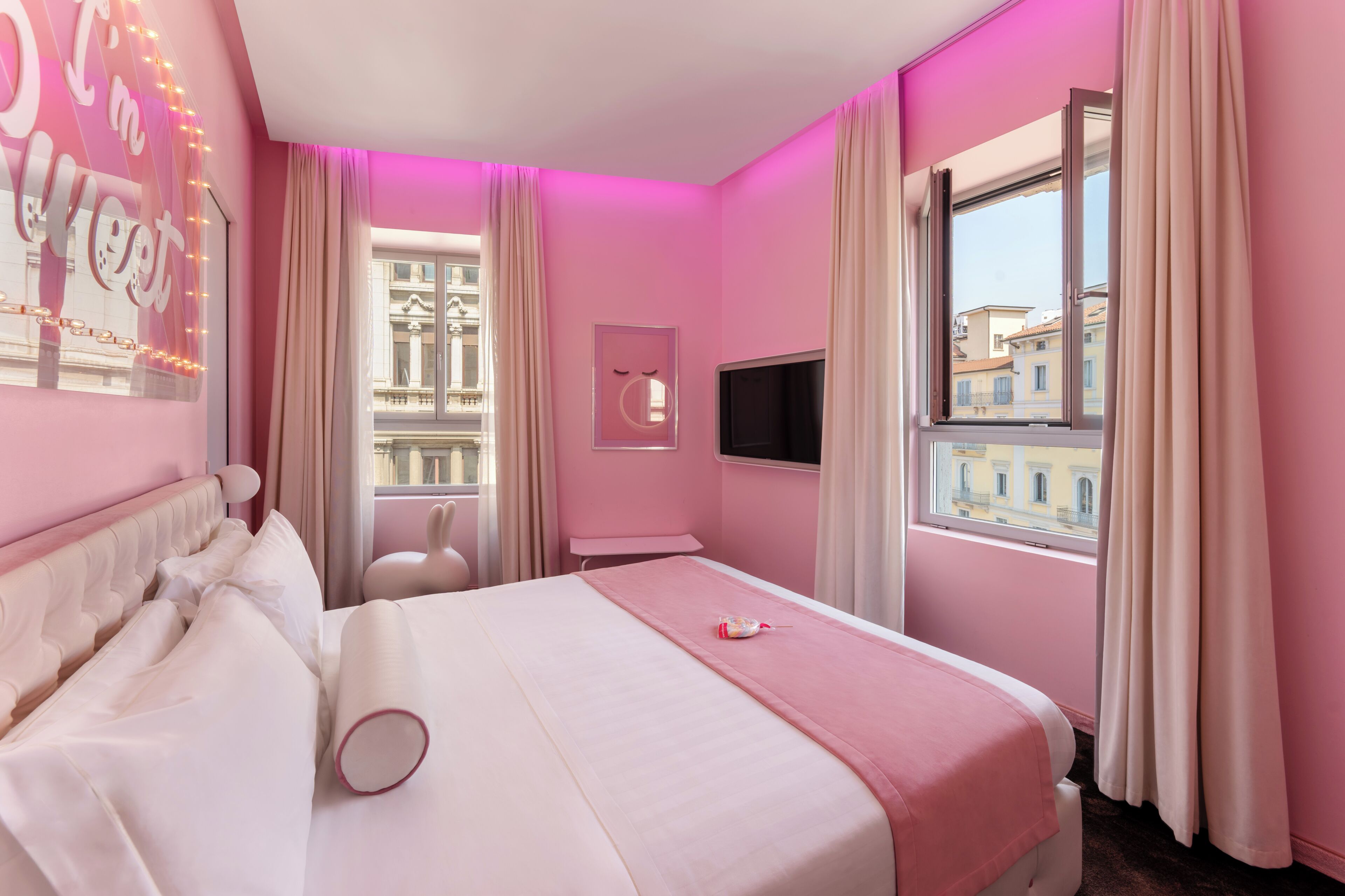 A pink-themed hotel room with a bed, a TV, a window, and a view of buildings.