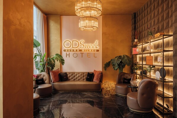 A hotel lobby with a sofa, chairs, a chandelier, and a wall with the hotel name. - ODSweet Duomo Milano Hotel (Milan)