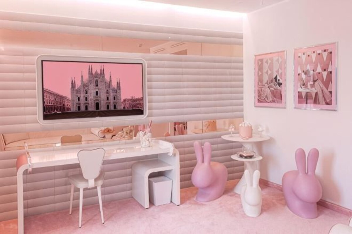 Une pièce avec un bureau blanc, des chaises, une sculpture de lapin rose et un téléviseur fixé au mur qui diffuse une image d’un bâtiment.