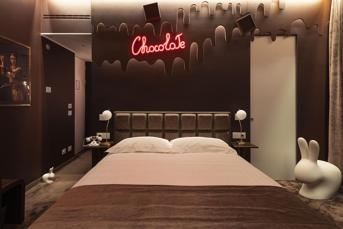 A modern hotel room with a large bed, a neon 'Chocolate' sign, a headboard, bedside tables, lamps, and a white rabbit sculpture.