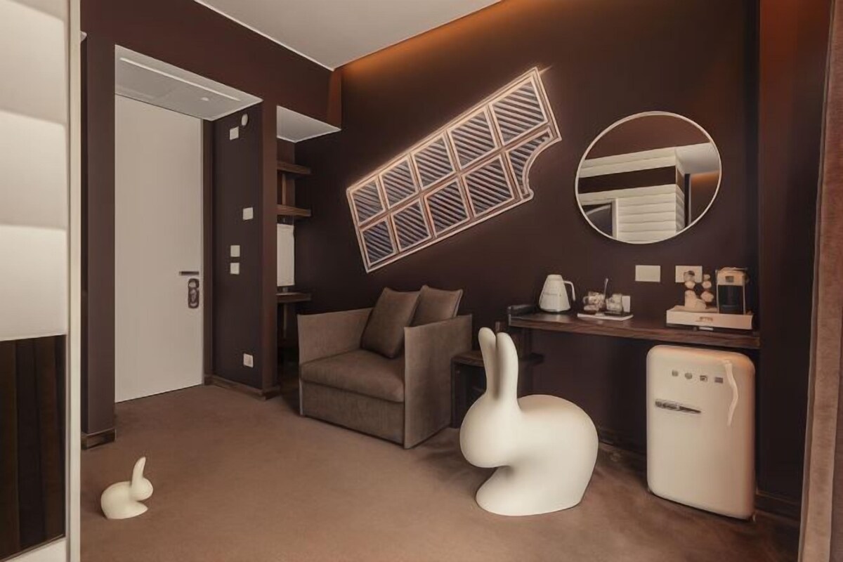 A modern hotel room with a sofa, a white rabbit sculpture, a refrigerator, and a wall-mounted mirror.
