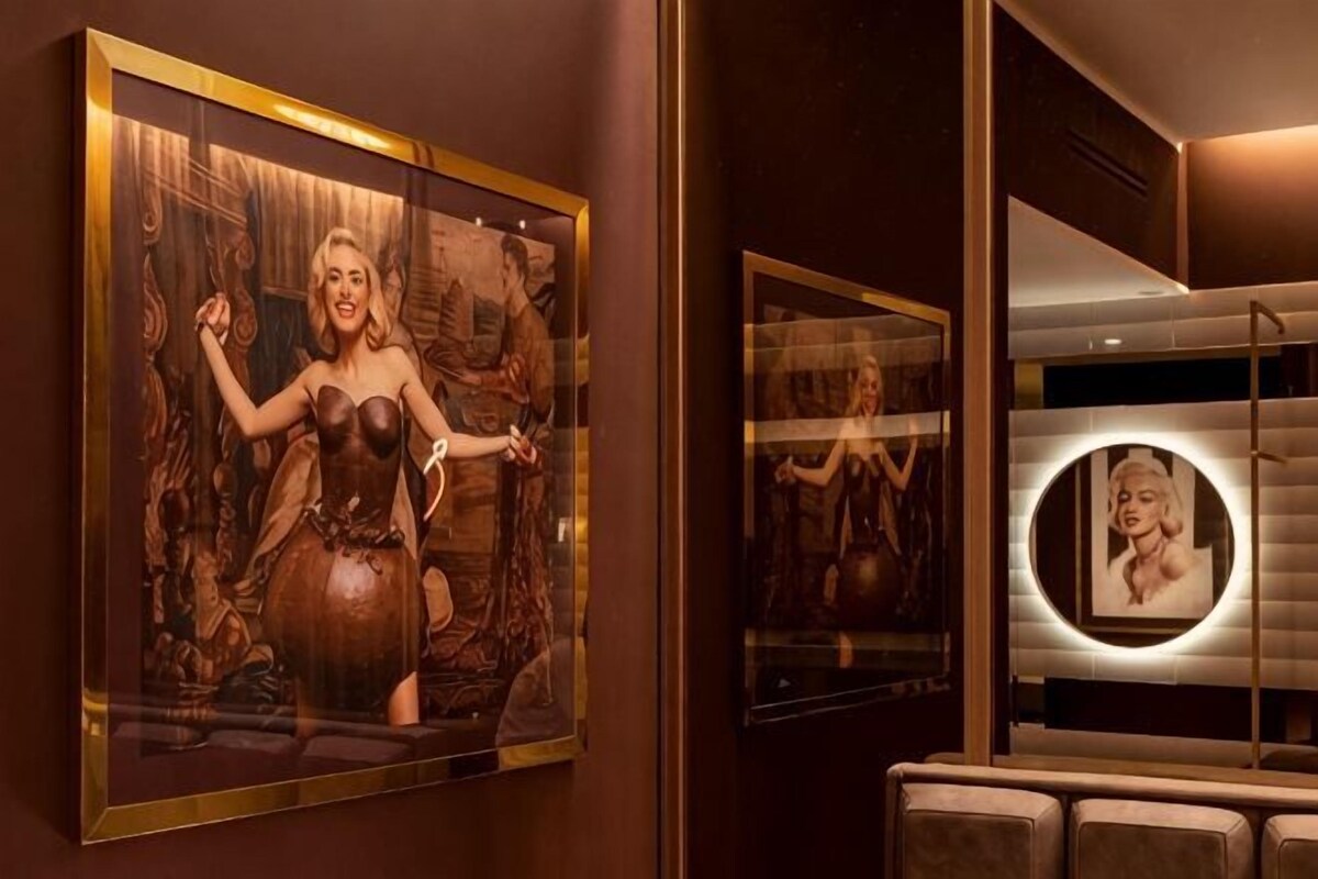 Un'opera d'arte incorniciata raffigura una donna con un vestito, uno specchio con cornice circolare e un'area salotto con poltrone imbottite.