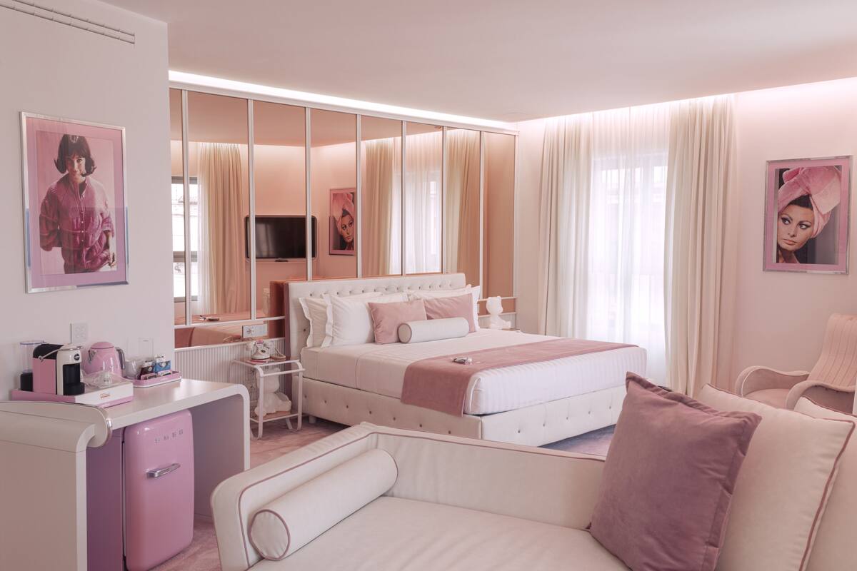 A modern bedroom with a large bed, bedside tables, a vanity, and a pink refrigerator.