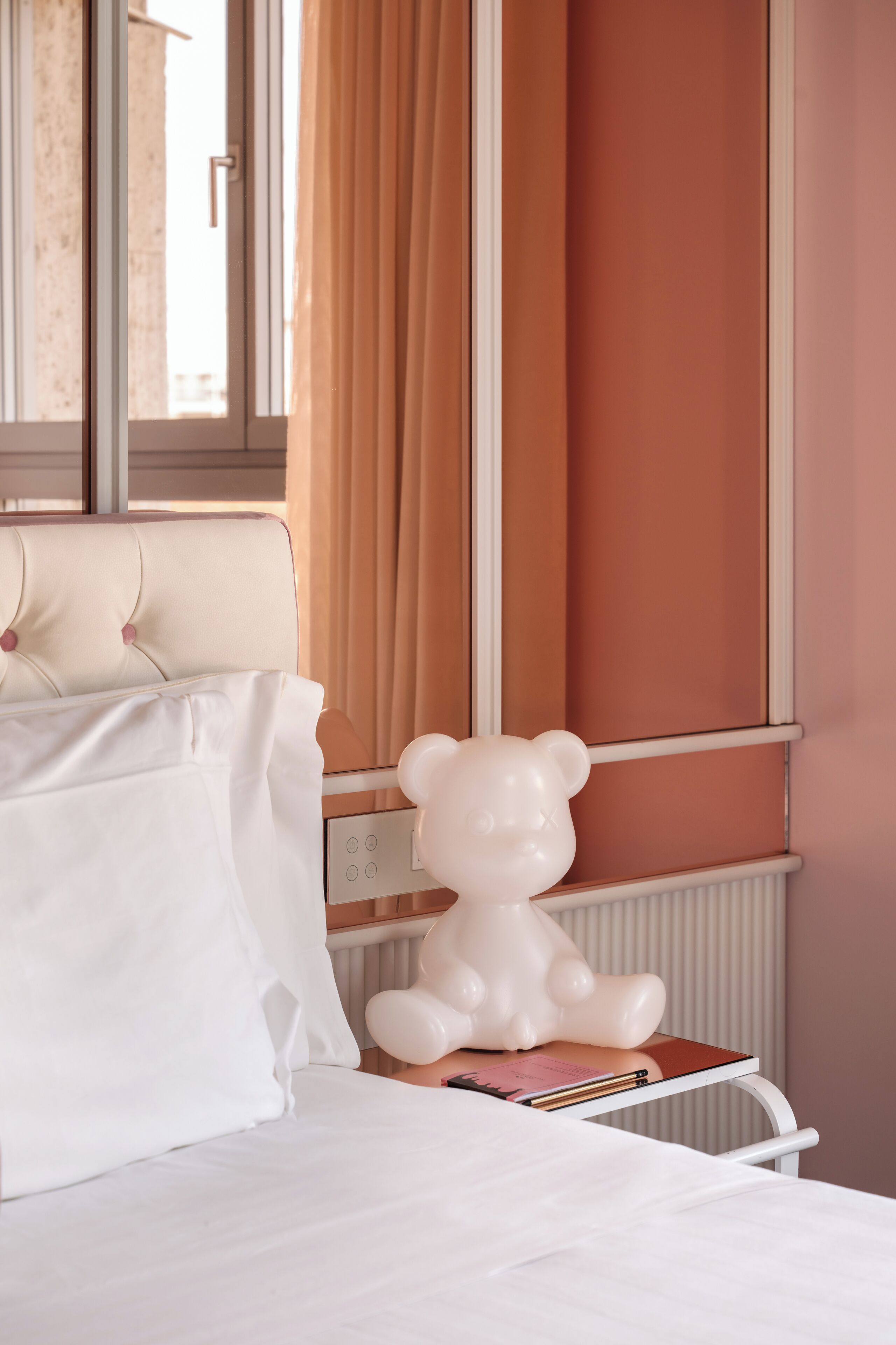 A hotel room with a bed, a white bear-shaped lamp, a window with curtains, and a radiator.