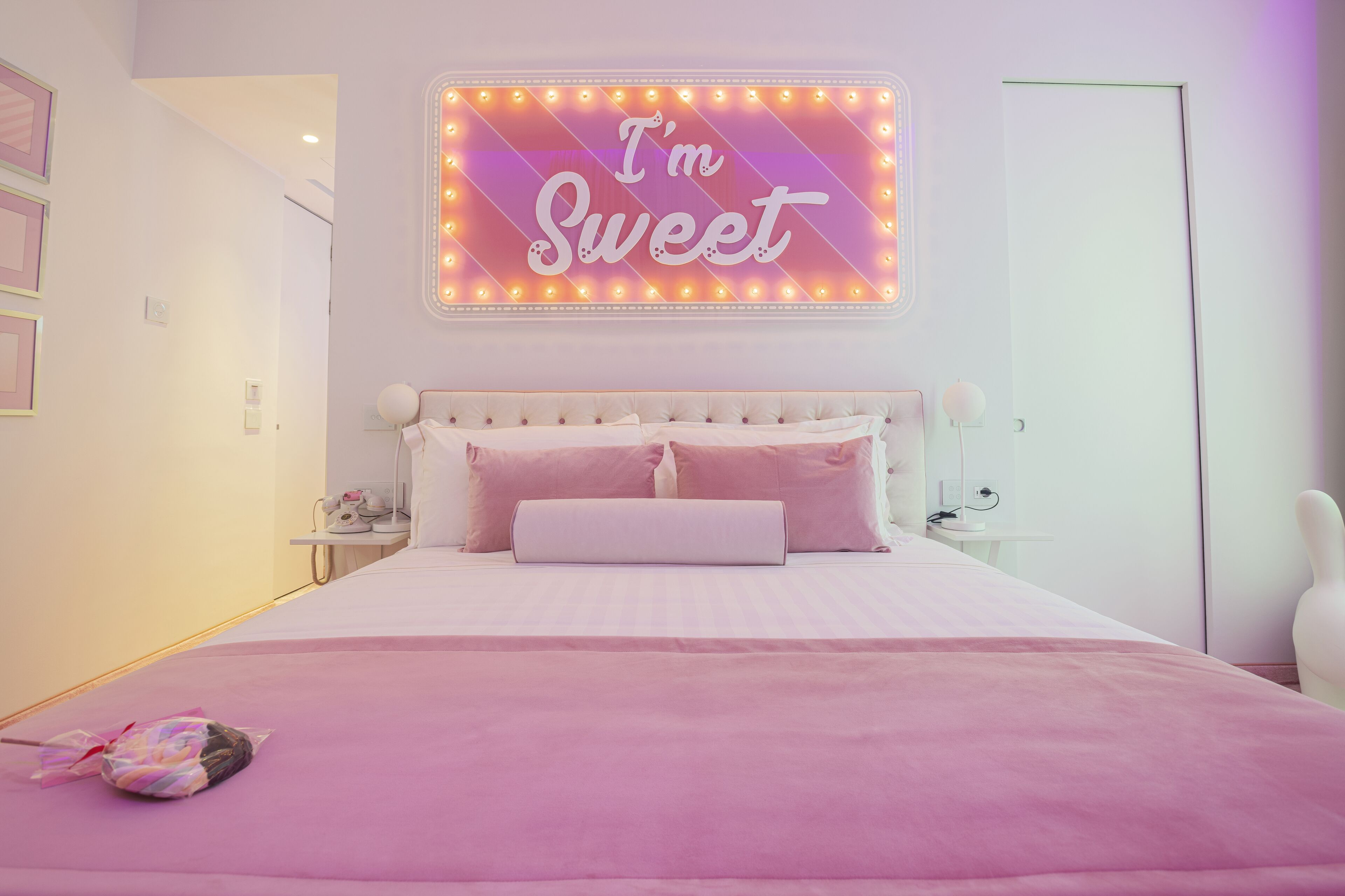 Una camera da letto con un letto, un neon con la scritta 'I'm Sweet' e una coperta rosa.