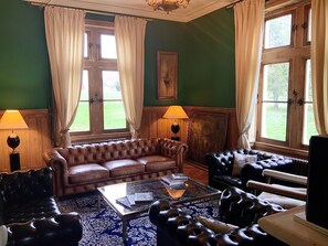 Una habitación con pared verde, dos ventanas con cortinas, un sofá de cuero marrón, un sillón de cuero negro y una mesa de centro de vidrio con libros.