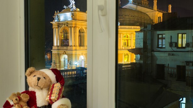 Un ours en peluche, vêtu d’un costume de Père Noël, est assis près d’une fenêtre donnant sur un bâtiment historique à l’architecture classique.