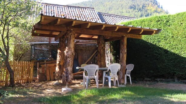A rustic outdoor seating area with a pergola, wooden chairs, and a stone wall in the background. - Family house in the center of Lozère (BALSIEGES)