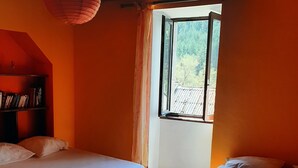 A room with two beds, wooden flooring, a ceiling with wooden beams, and a window with curtains. - Family house in the center of Lozère (BALSIEGES)