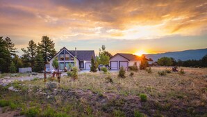 A modern house with a large garage and a deck, surrounded by a natural landscape with trees and a sunset in the background. - Ideal Mountain Retreat!   Peak Views, 15 Minutes to Salida and BV.   Fast WIFI. (Nathrop)