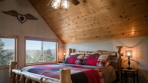 A log cabin bedroom with a large bed, wooden headboard, and a view of the outdoors through two windows. - Ideal Mountain Retreat!   Peak Views, 15 Minutes to Salida and BV.   Fast WIFI. (Nathrop)
