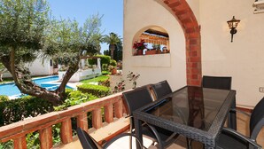 Un patio con mesa y sillas de vidrio, con vista a una piscina y abundante follaje.