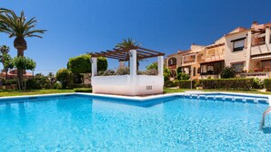 Una piscina con una pérgola, rodeada de césped cuidado y palmeras.
