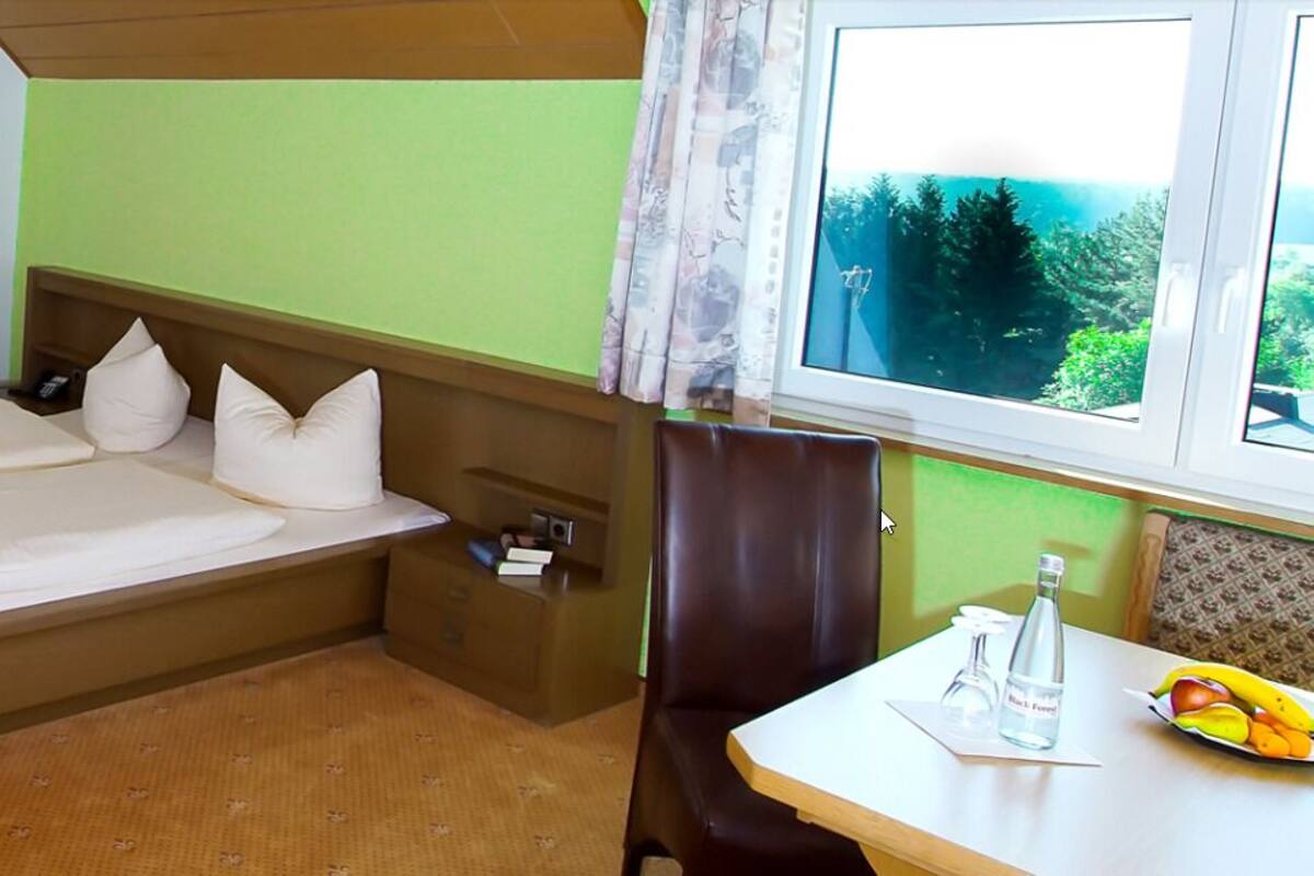 Une chambre d’hôtel avec un lit, un bureau, une lampe, une fenêtre donnant sur des arbres et une table avec une bouteille et des fruits.