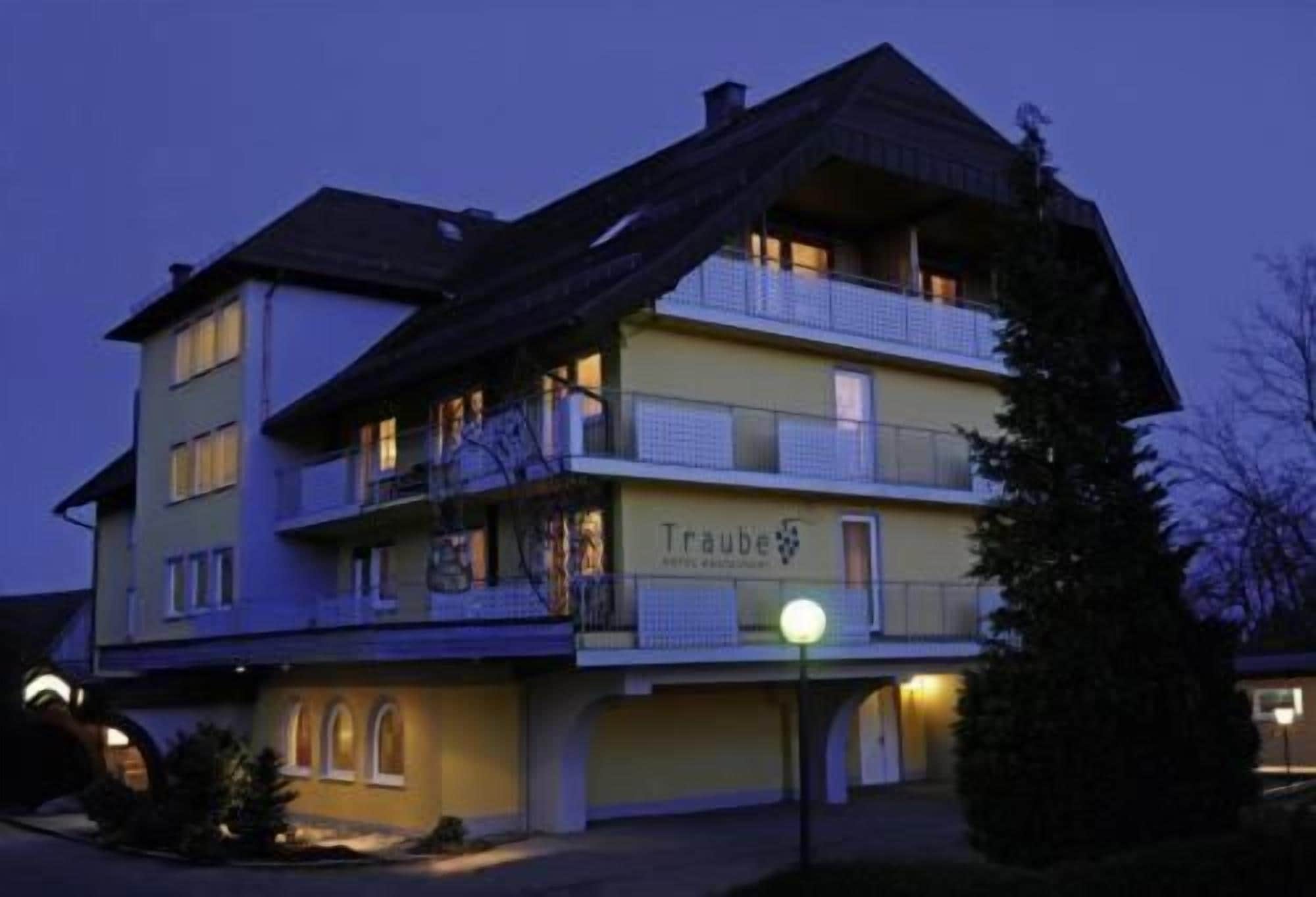 Un hôtel à plusieurs étages, doté d’un balcon au deuxième étage, d’une enseigne indiquant « Traube » et d’une entrée bien éclairée.