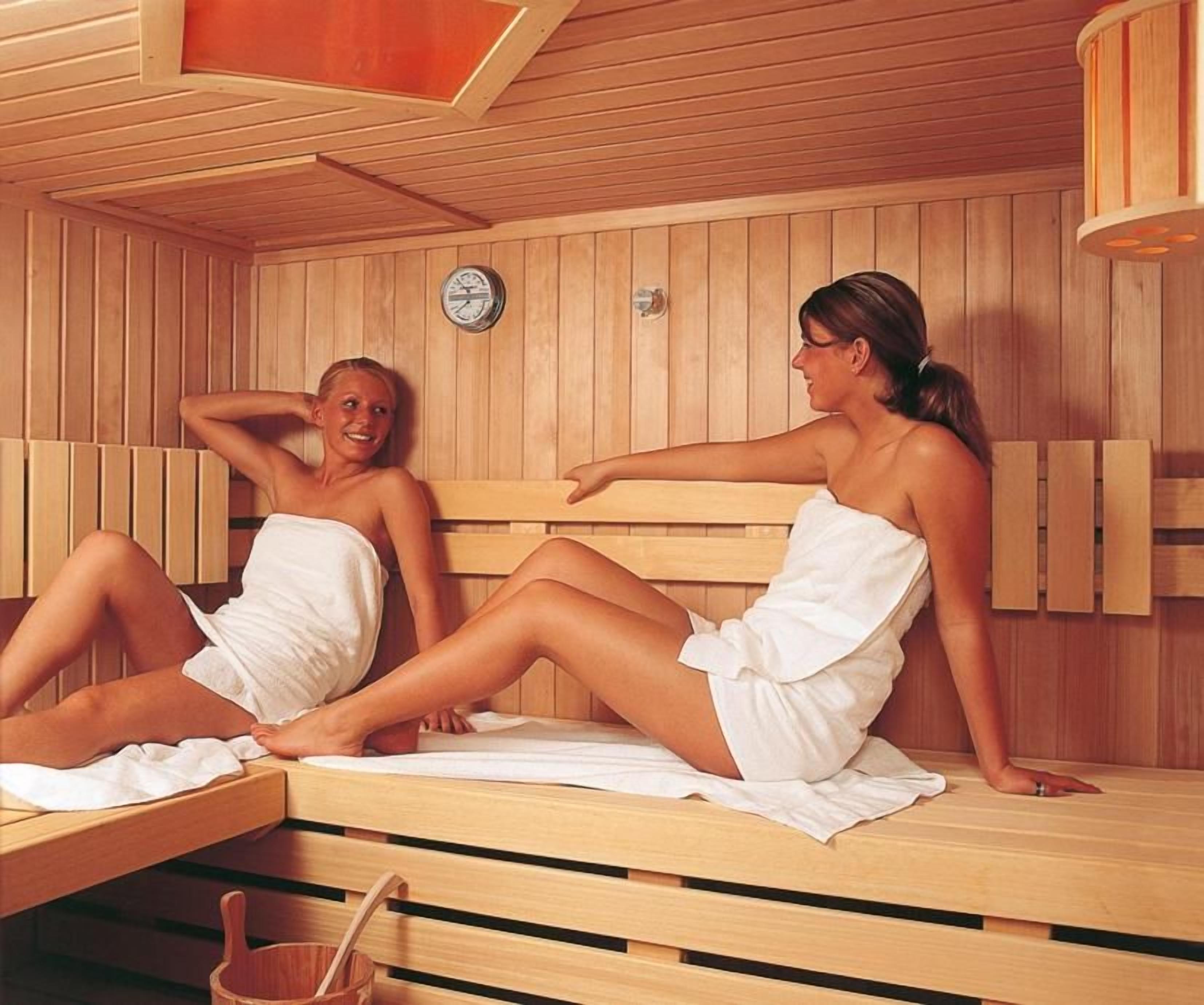 Twee personen in een sauna met houten banken en een zichtbare klok aan de muur.