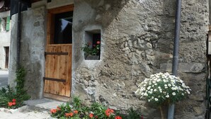 A rustic stone building with a wooden door and windows, surrounded by a flower bed with red and white flowers.