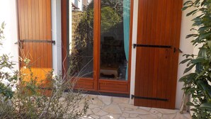 A wooden door with a glass panel, flanked by two wooden shutters, set in a stone-paved entrance area with greenery around. - Charming T2 house in a quiet condominium near the beaches. (Vaux-sur-Mer)