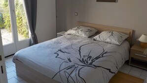 A bedroom with a bed, a nightstand, a lamp, and a painting on the wall. - Charming T2 house in a quiet condominium near the beaches. (Vaux-sur-Mer)