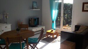 A living room with a wooden dining table, teal cushioned chairs, a black sofa, and a television on a white cabinet. - Charming T2 house in a quiet condominium near the beaches. (Vaux-sur-Mer)